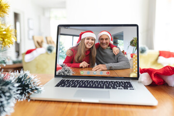 Caucasian father and adult daughter in santa hats smiling on laptop video call screen at christmas