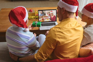 Caucasian father and sons in santa hats making laptop christmas video call with family at table