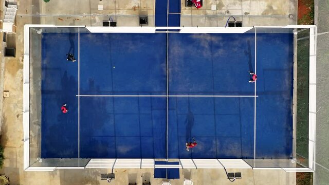 View from above, aerial footage, slow motion video of some people playing on a blue padel court. Padel is a mix between Tennis and Squash. It's usually played in doubles on an enclosed court.