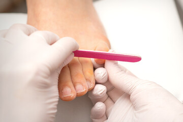 Pedicurist hands in protective rubber gloves filing toenails on feet with a nail file in a beauty salon