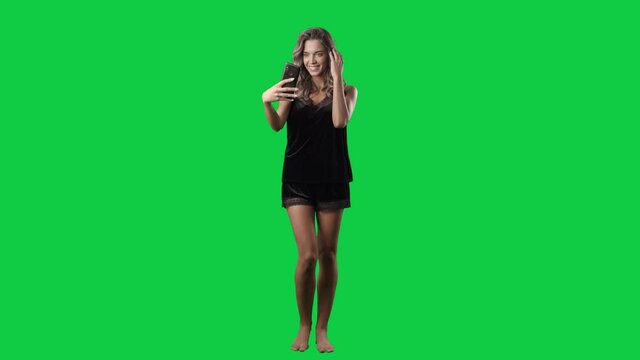 Cute young woman in black satin nightwear taking selfies puckering lips. Full body isolated on green screen background