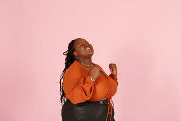 Excited young black plus size body positive woman with dreadlocks in orange top happy of big win poses for camera on pink background in studio closeup