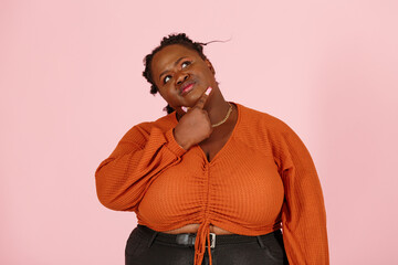Thoughtful young black overweight body positive woman with dreadlocks in orange top touches chin looking up on light pink background in studio closeup - Powered by Adobe