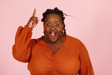 Funny surprised young black overweight body positive woman with dreadlocks in orange top gets cool idea looking at camera on light pink background in studio closeup