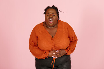 Young black plus size body positive woman patient in orange top suffers from stomach ache standing on light pink background in studio waist up