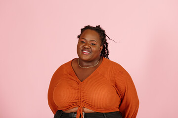Shy young black plus size body positive woman with dreadlocks in orange top smiles to camera on pink background in studio