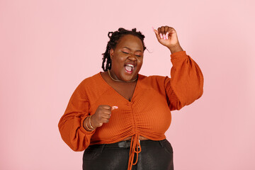 Excited young black plus size body positive woman with dreadlocks in orange top happy of big win poses for camera on pink background in studio closeup - 469267031