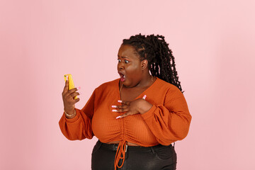 Wondered young black plus size body positive woman with dreadlocks in orange top holds smartphone standing on light pink background in studio closeup