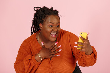 Cheerful young black plus size body positive woman with dreadlocks in orange top talks by videochat holding modern smartphone on pink background in studio closeup