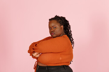 Critical young black plus size body positive woman with dreadlocks in orange top looks at camera crossing arms on pink background in studio