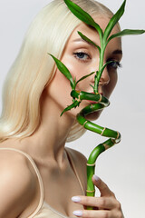 A blonde-haired blue-eyed young woman with smooth fresh skin holds a green bamboo branch near her face. Advertising of organic cosmetics. Skin and hair care products with natural composition.