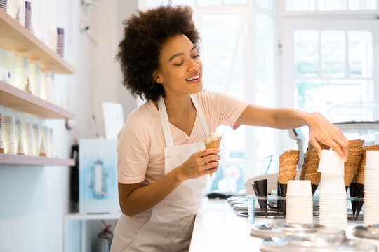 A beautiful mixed-race woman is smiling behind the stand of an ice cream parlor and serving the ice cream in the cones.