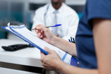 Closeup of patient mother writing medical information on document discussing sickness symptoms with african american doctor during clinical appointment in hospital office. Health care service