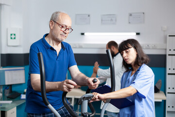 Aged patient doing physical workout for injury recovery with medical assistant. Man using...