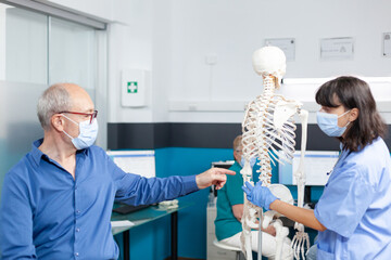 Obraz premium Patient and nurse with face masks looking at spine bones on human skeleton for osteopathy examination and physical recovery. Assistant showing spinal cord to old man during pandemic