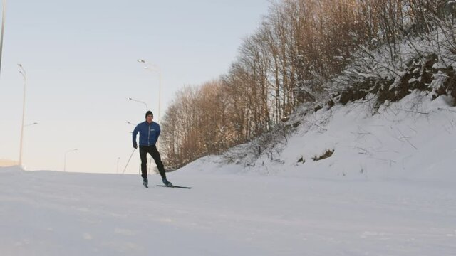 Low angle of healthy Caucasian man wearing sporty outfit, skiing downhill from distance on white snow by forest, stopping before camera