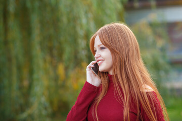 Portrait casual of a Red hair girl in a red dress, looking a side and smiles, speaking on a phone. stays outdoor. model beautiful, long straight hair, hands and arms. Blurred details