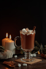 Hot cocoa or chocolate with melting marshmallows in a tall glass. Burning candles , a blanket and fir branches in the background