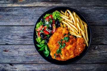 Breaded fried pork chop with French fries and fresh vegetables on wooden table
