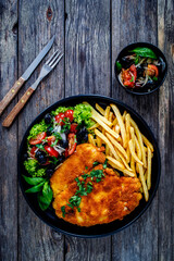 Breaded fried pork chop with French fries and fresh vegetables on wooden table
