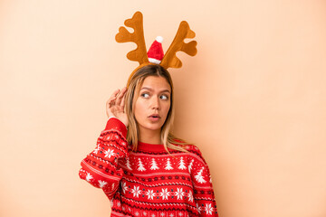 Young caucasian woman wearing a christmas reindeer hat isolated on beige background trying to listening a gossip. © Asier