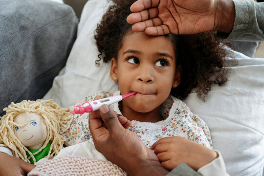 Father with thermometer measuring temperature of daughter on sofa