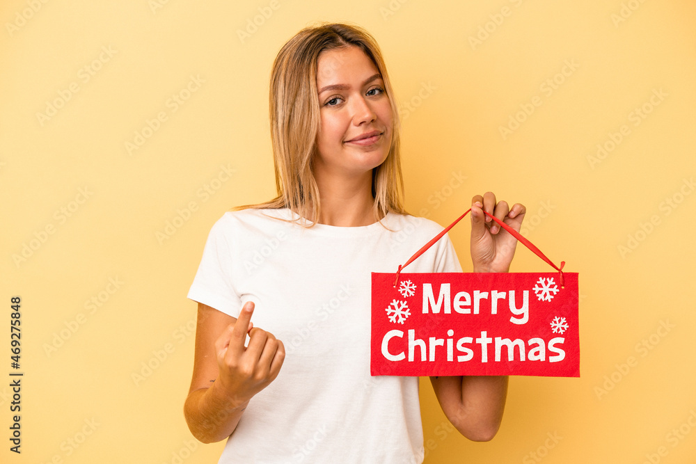 Wall mural Young caucasian woman holding a merry christmas placard isolated on yellow background pointing with finger at you as if inviting come closer.