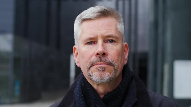 Close-up portrait of serious and strict looking middle aged old man in city . Push-in on elderly senior citizen downtown looking at camera with neutral expression. People and urban lifestyle.
