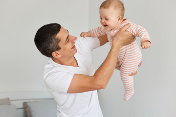 Indoor shot of brunette man wearing white casual t shirt holding his infant daughter, throwing baby...