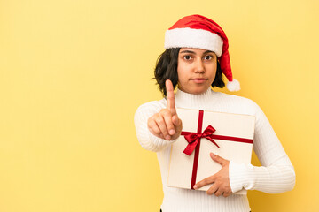 Young latin woman celebrating christmas holding a gift isolated on yellow background showing number one with finger.