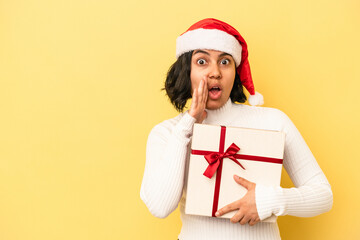 Young latin woman celebrating christmas holding a gift isolated on yellow background is saying a secret hot braking news and looking aside