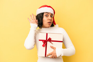 Young latin woman celebrating christmas holding a gift isolated on yellow background trying to listening a gossip.
