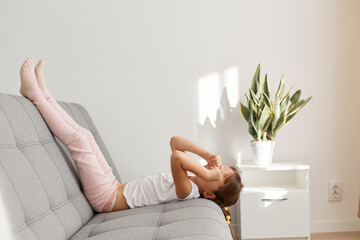 Sleepy adorable child lying on gray comfortable sofa with raised legs and rubbing her eyes, feels tired, wants sleeping , needs time to rest, wearing casual style clothing.