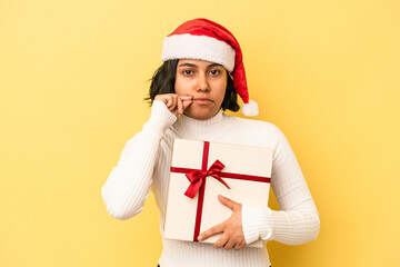 Young latin woman celebrating christmas holding a gift isolated on yellow background with fingers on lips keeping a secret.