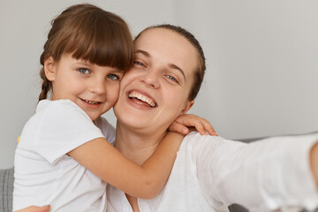 Extremely happy smiling female taking selfie with charming dark haired daughter, looking smiling at cell phone camera and hugging point of view photo, family posing at home.