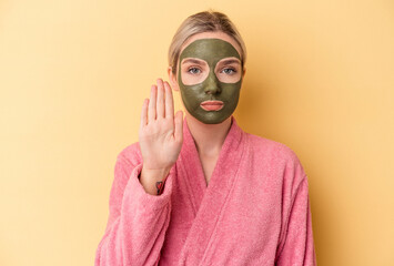 Young caucasian woman wearing face mask isolated on yellow background standing with outstretched hand showing stop sign, preventing you.