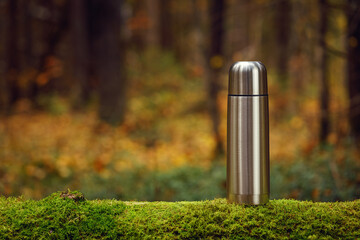 Steel thermos with delicious hot tea on an old mossy tree in focus. The background of the autumn...