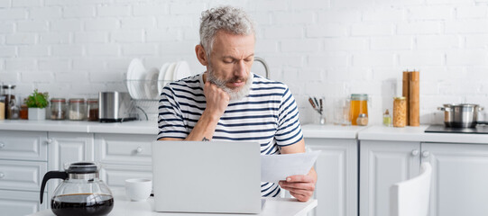 Mature man in striped t-shirt holding paper near laptop and coffee pot in kitchen, banner.