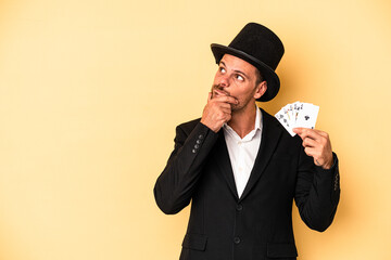 Young caucasian wizard holding magic card isolated on yellow background looking sideways with doubtful and skeptical expression.