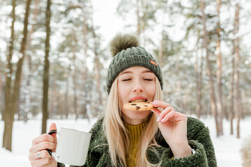 Beautiful young girl enjoying winter outdoors. Pretty women having fun with snow in forest or park. Winter picnic with cocoa and cookies.