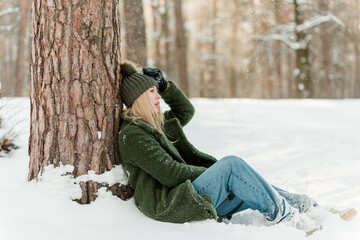 Fototapeta na wymiar Beautiful young girl is sitting near tree enjoying winter outdoors. Pretty women having fun with snow in forest
