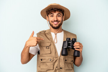 Young mixed race man holding binoculars isolated on blue background person pointing by hand to a shirt copy space, proud and confident