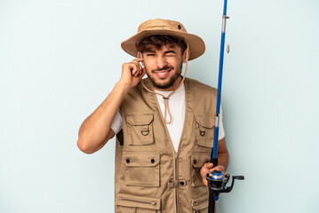 Young mixed race fisherman holding a rod isolated on blue background covering ears with hands.