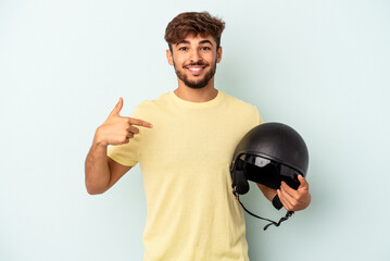 Naklejka premium Young mixed race man holding motorcycle helmet isolated on blue background person pointing by hand to a shirt copy space, proud and confident