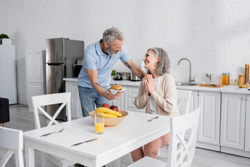 Obraz premium Man holding plate with pancakes while smiling wife showing please gesture in kitchen.