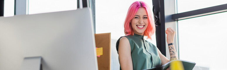 cheerful businesswoman with pink hair holding folder and looking at computer monitor, banner.
