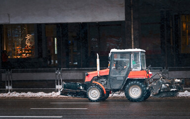 Fototapeta na wymiar Small tractor with rotary brush and snowplow driving on city road on snowy winter day. Municipal city service cleaning snow , tractor with rotating brush clears road from ice and snow.