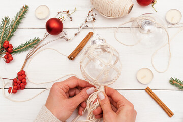 Fototapeta na wymiar A woman decorates a transparent Christmas ball with macrame-style weaving,handmade decor in eco-style,hands close-up,top view.Christmas, New Year and eco-friendly concept.