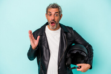 Middle age biker caucasian man holding helmet isolated on blue background  surprised and shocked.