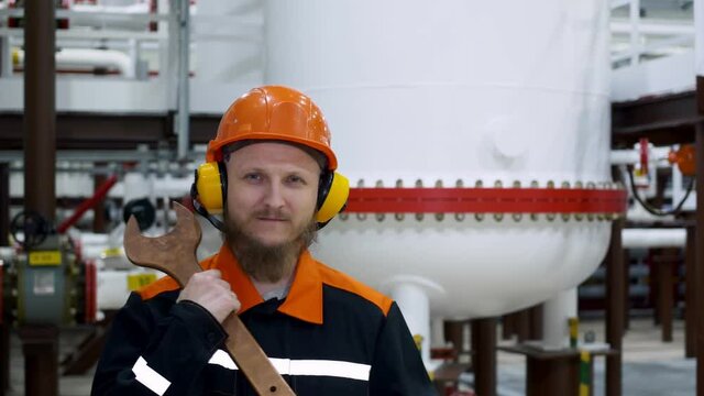 An oil and gas worker stands in a hard hat and ear protectors in a gas liquefaction shop, behind him is a container of gas, and in his hands is a large wrench. Workplace repairman.